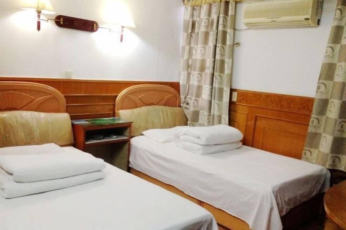 A hotel room with two single beds, a wooden headboard, a nightstand, a lamp, and a wall-mounted air conditioner.