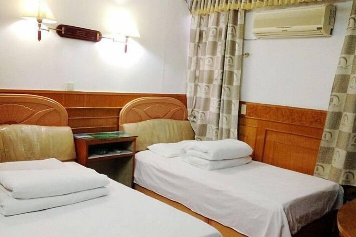 A hotel room with two single beds, a nightstand, a lamp, an air conditioner, and a patterned curtain.