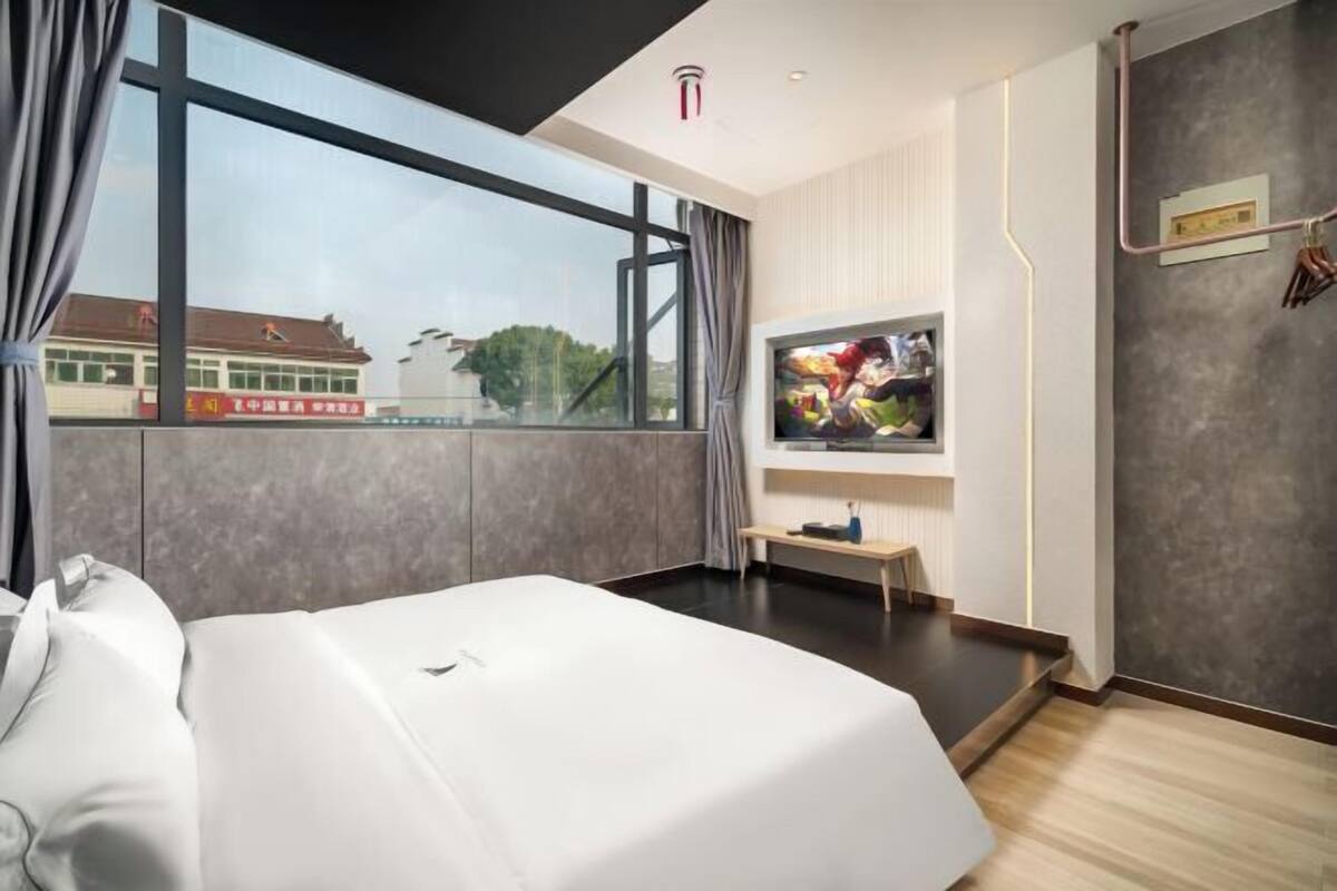 A modern hotel room with a large bed, a TV mounted on the wall, and a view of the outside through a large window.