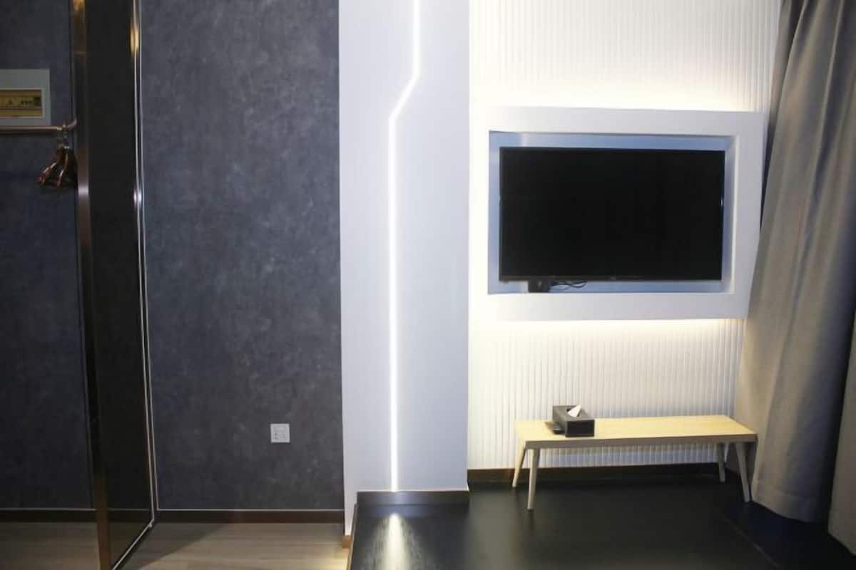 A modern hotel room with a large flat-screen TV mounted on the wall, a small wooden bench, and a glass door leading to a shower area.