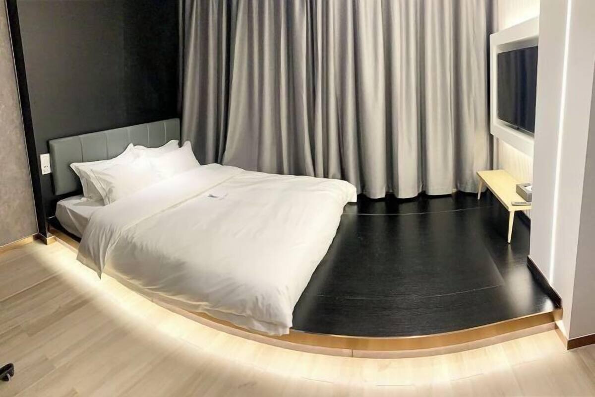 A modern bedroom with a curved bed, grey curtains, a small bedside table, and a TV mounted on the wall.