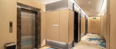 A hotel hallway with elevators, carpeted flooring, and a wall-mounted light fixture.