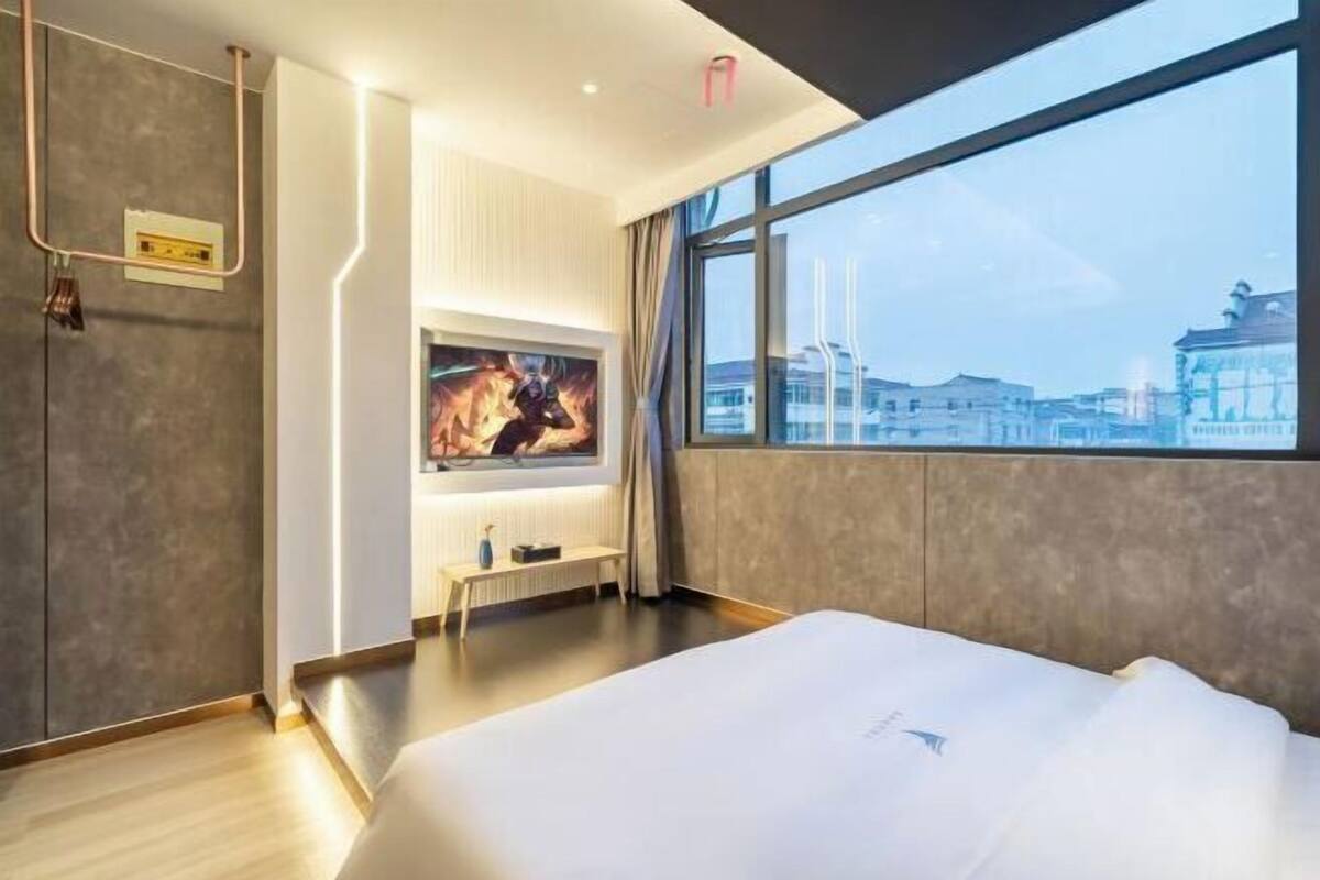 A hotel room with a large bed, a television mounted on the wall, a window with a city view, and a wooden bench.