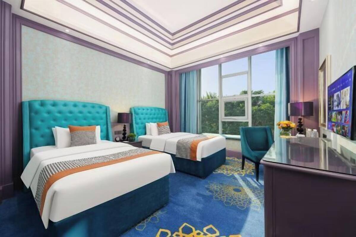 A hotel room with two beds, a desk, and a large window offering a view of greenery.