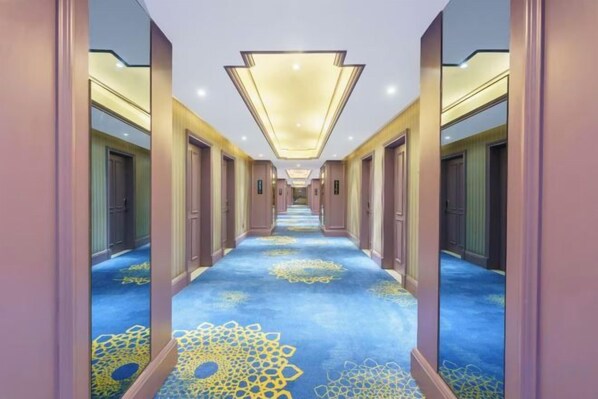 Pasillo de hotel con paredes espejadas, alfombra estampada y lámpara en el techo.