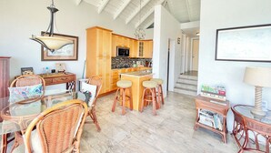 Una cocina amplia con gabinetes de madera y una isla central, un comedor con sillas de mimbre y una mesa de vidrio, y un pasillo que conduce a otra habitación.