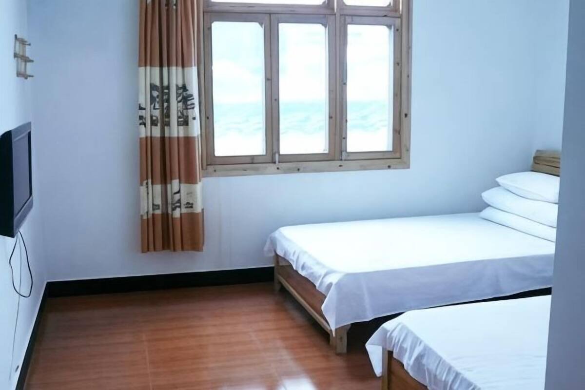 A room with a wooden floor, a bed with white bedding, a window with curtains, and a television on the wall.