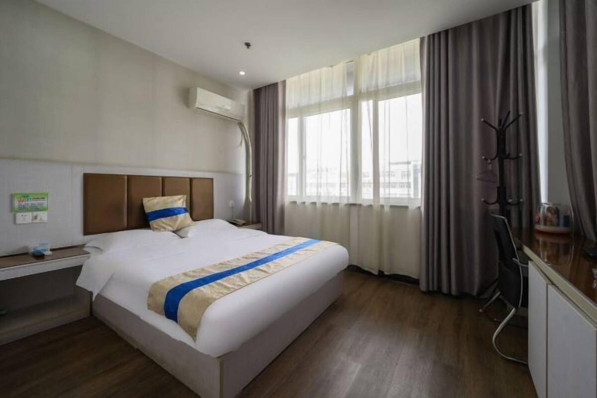 A hotel room with a bed, a desk, a chair, a coat rack, and a window with curtains.