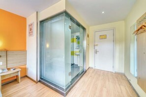 A hotel room with a glass partition, wooden flooring, a bench, and a door with a light switch.