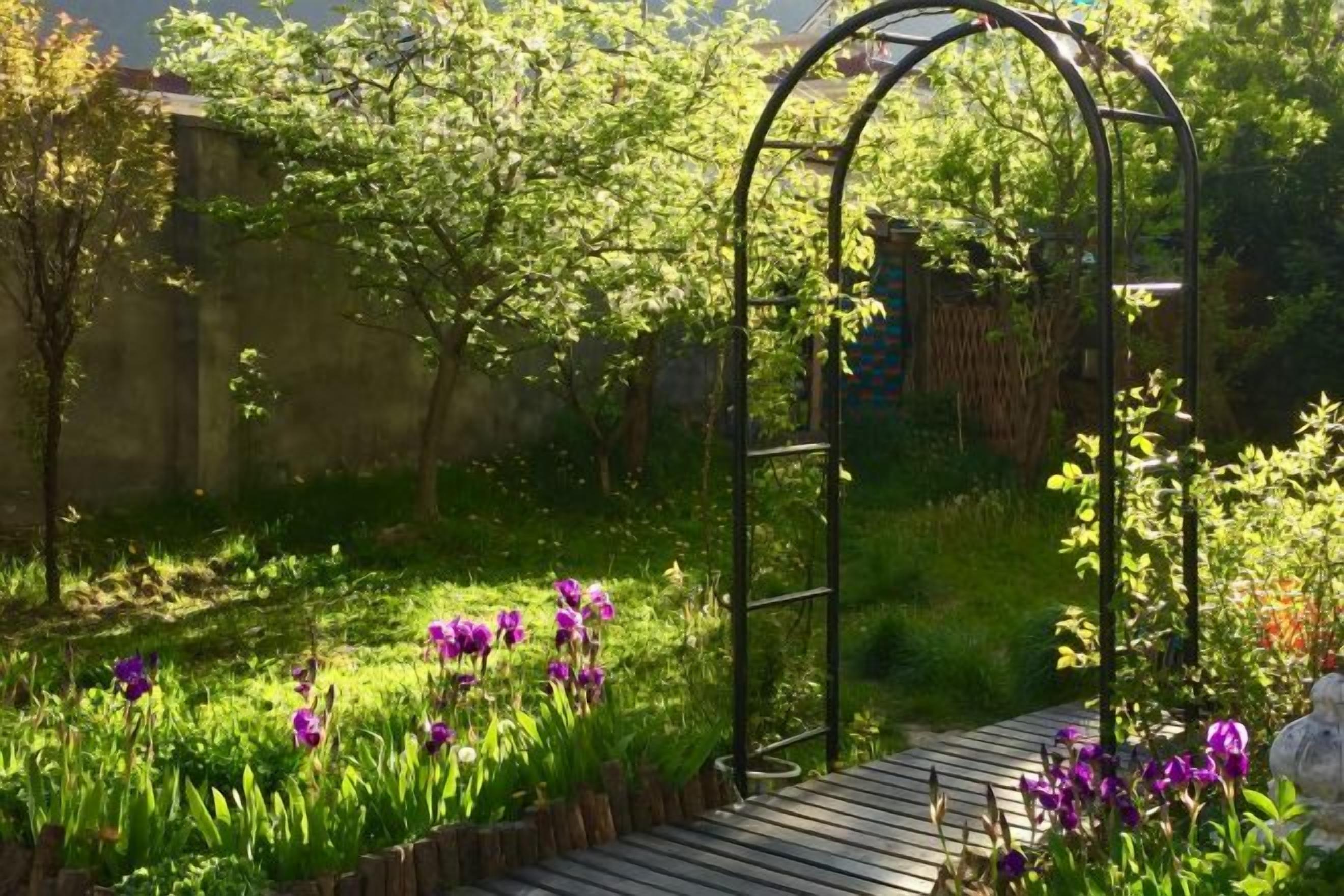 A garden with a wooden pathway, a metal archway, and various plants and flowers.
