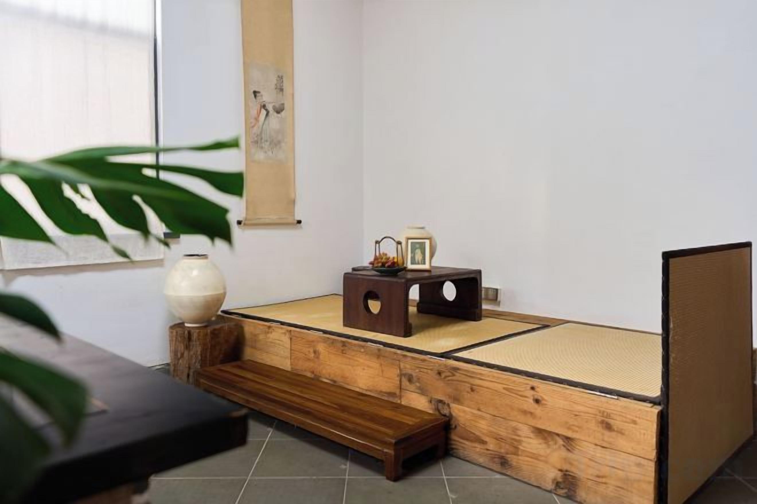 A tatami mat room with a low wooden bench, a small table with a vase and a picture frame, and a large plant.