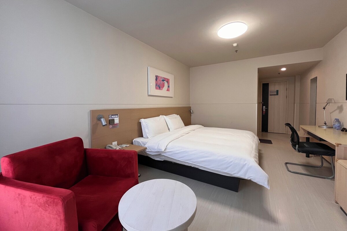 Ein Hotelzimmer mit einem Bett, einem roten Sessel, einem Schreibtisch mit Stuhl und einem kleinen Tisch.