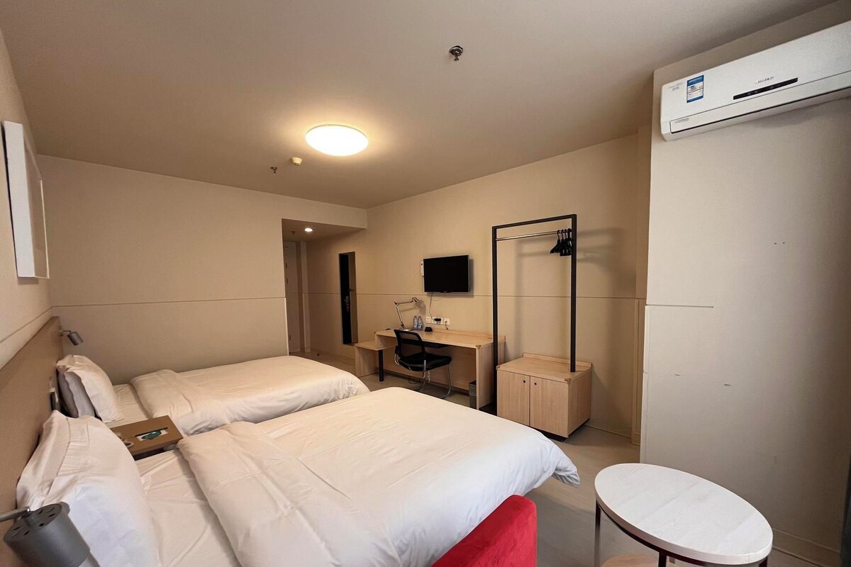 Ein Hotelzimmer mit zwei Betten, einem Schreibtisch mit Stuhl, einem kleinen Tisch, einem Spiegel und einer Klimaanlage.