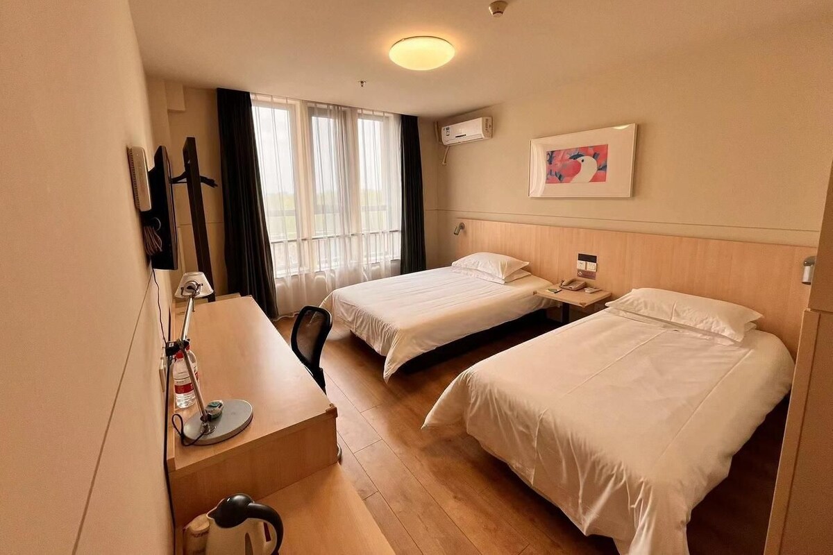 Ein Hotelzimmer mit zwei Betten, einem Schreibtisch, einem Stuhl, einem Wasserkocher und einem Fenster mit Vorhängen.