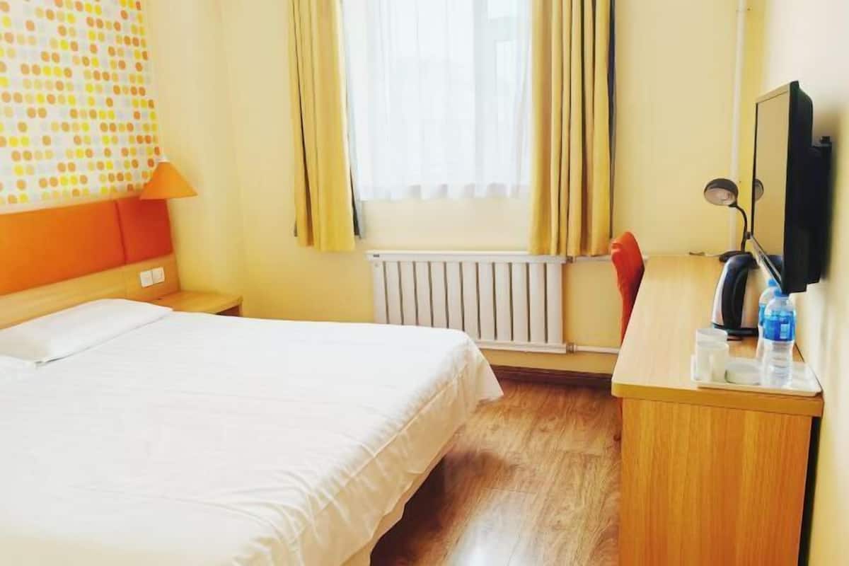 A hotel room with a bed, a desk with a computer, a window with curtains, and a radiator.