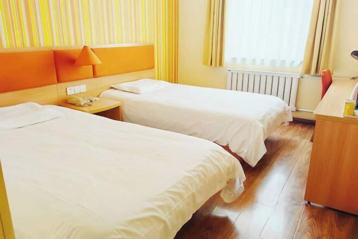 A hotel room with two single beds, a wooden headboard, a bedside table with a lamp, and a radiator.