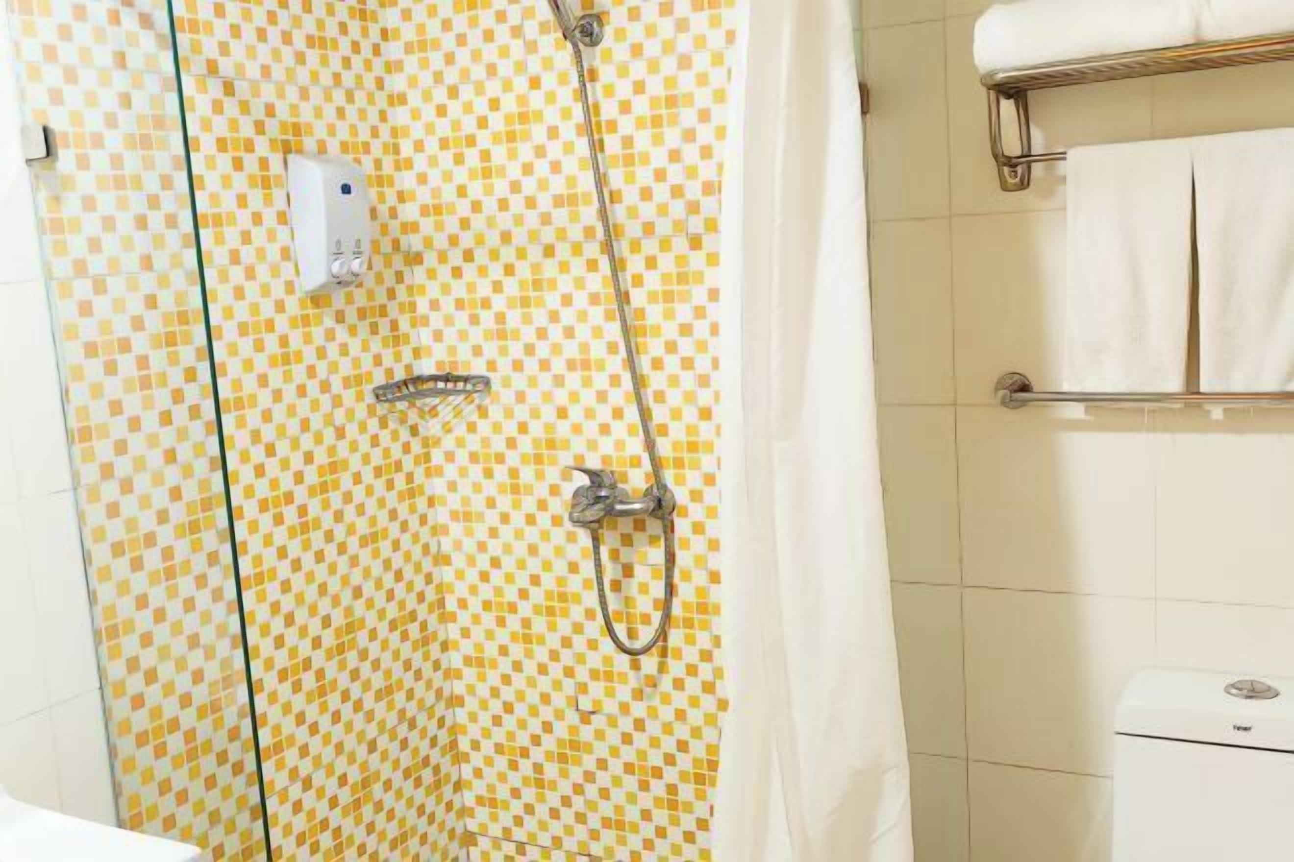 Un baño con pared de azulejos a cuadros, ducha con cabezal de ducha de mano, toallero con toallas blancas y un inodoro con etiqueta de marca visible.