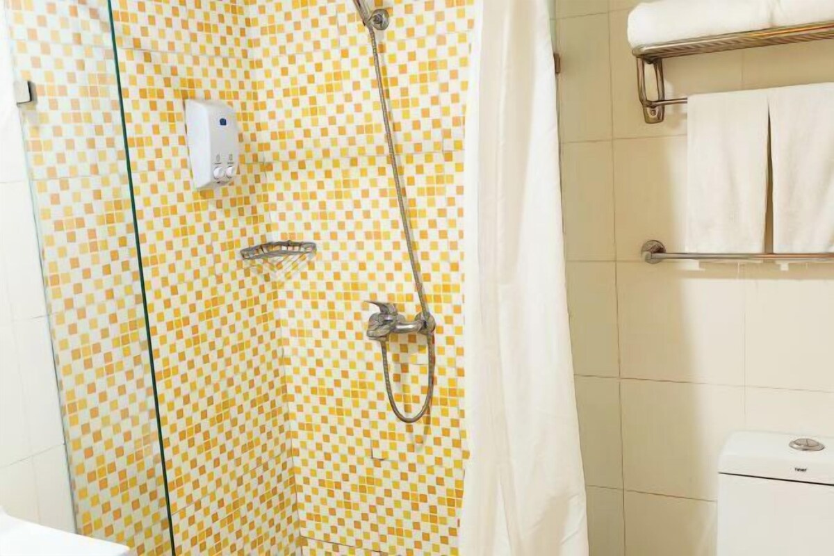 A bathroom with a checkered tile wall, a shower with a handheld showerhead, a towel rack with white towels, and a toilet with a visible brand label.