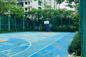 Een basketbalveld buiten, omringd door een groen hek en bomen, met een gebouw op de achtergrond.