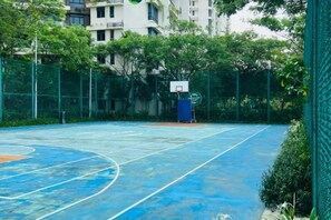 Ein Basketballplatz im Freien mit blauer Spielfläche, umgeben von einem grünen Zaun und Bäumen.