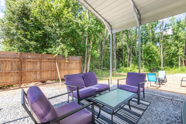 An outdoor patio with a covered seating area, including a sofa and chairs, surrounded by a wooden fence and trees. - Iconic Airstream Camper, PRIVATE Pet Friendly, Near Sportplex, 8 mi to Beach. (Foley)