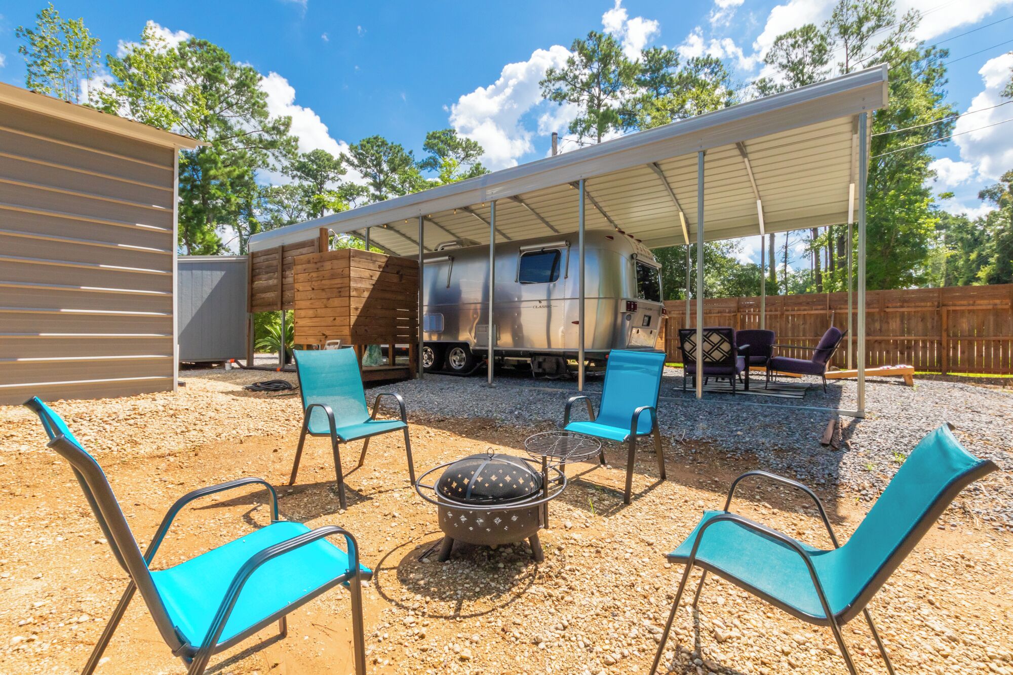 Un espacio de estar al aire libre cubierto con cuatro sillas azules, un brasero, una caravana y una estructura de madera.