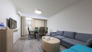 A modern hotel room with a grey sofa, a blue ottoman, a wooden coffee table, a TV on a stand, a desk with chairs, and a window with curtains. - HanTing Hotel (Yueyanglou District Pipawang Overpass) (Yueyang)