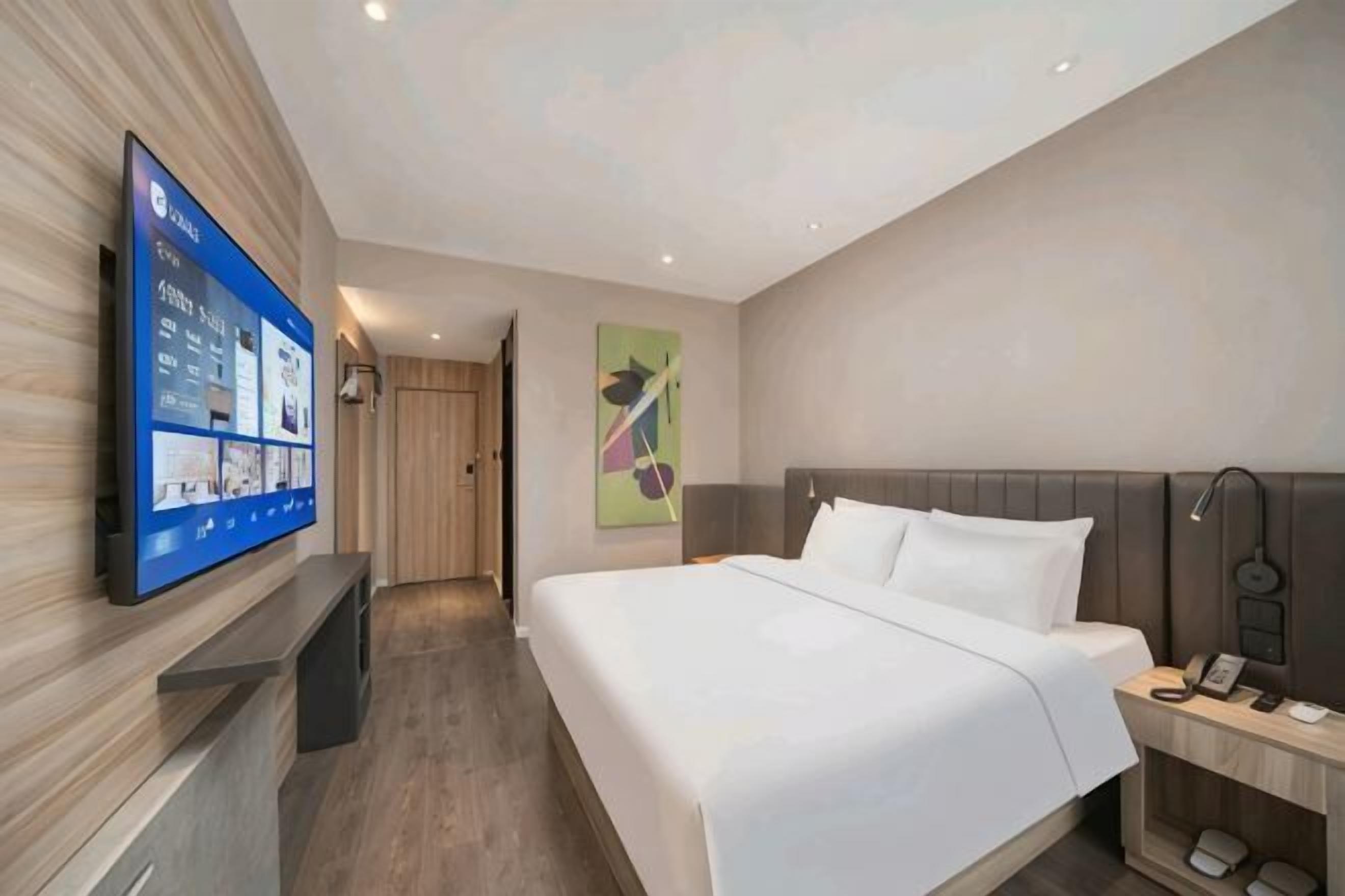 Une chambre d’hôtel moderne dotée d’une grande télévision à écran plat, d’un lit avec du linge de maison blanc, d’une porte en bois et d’une lampe fixée au mur.