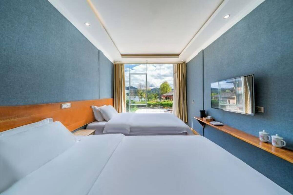 Une chambre d’hôtel avec deux lits, une télévision, une fenêtre donnant sur un espace verdoyant et une étagère en bois avec quelques objets.