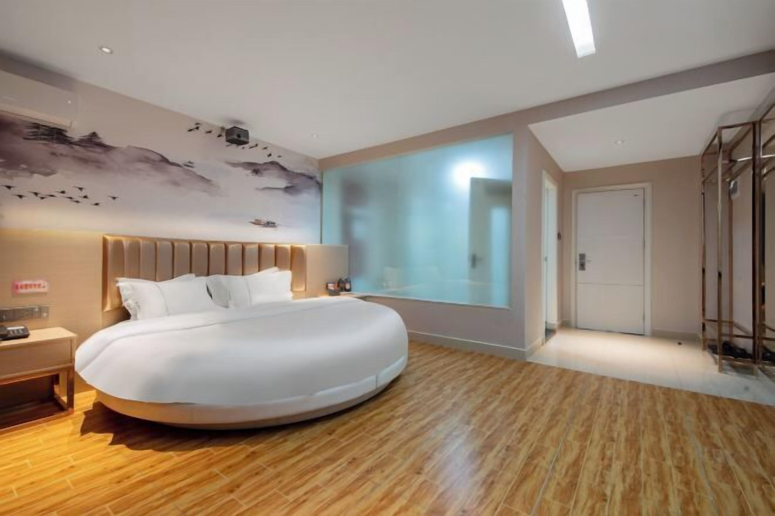 A modern hotel room with a large bed, a nightstand, a wall-mounted lamp, and a large window.