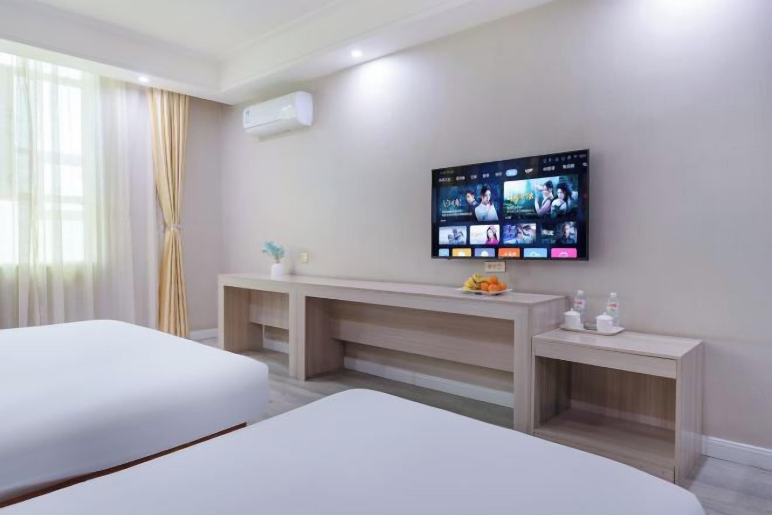 Habitación de hotel con dos camas, un televisor instalado en la pared mostrando varios canales, una mesita con un frutero y una mesita de noche con artículos de aseo.
