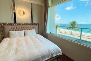 A bed with a tufted headboard, a large window with a view of a beach and palm trees, and a wall-mounted light fixture. - Angel Inn (Hualien)