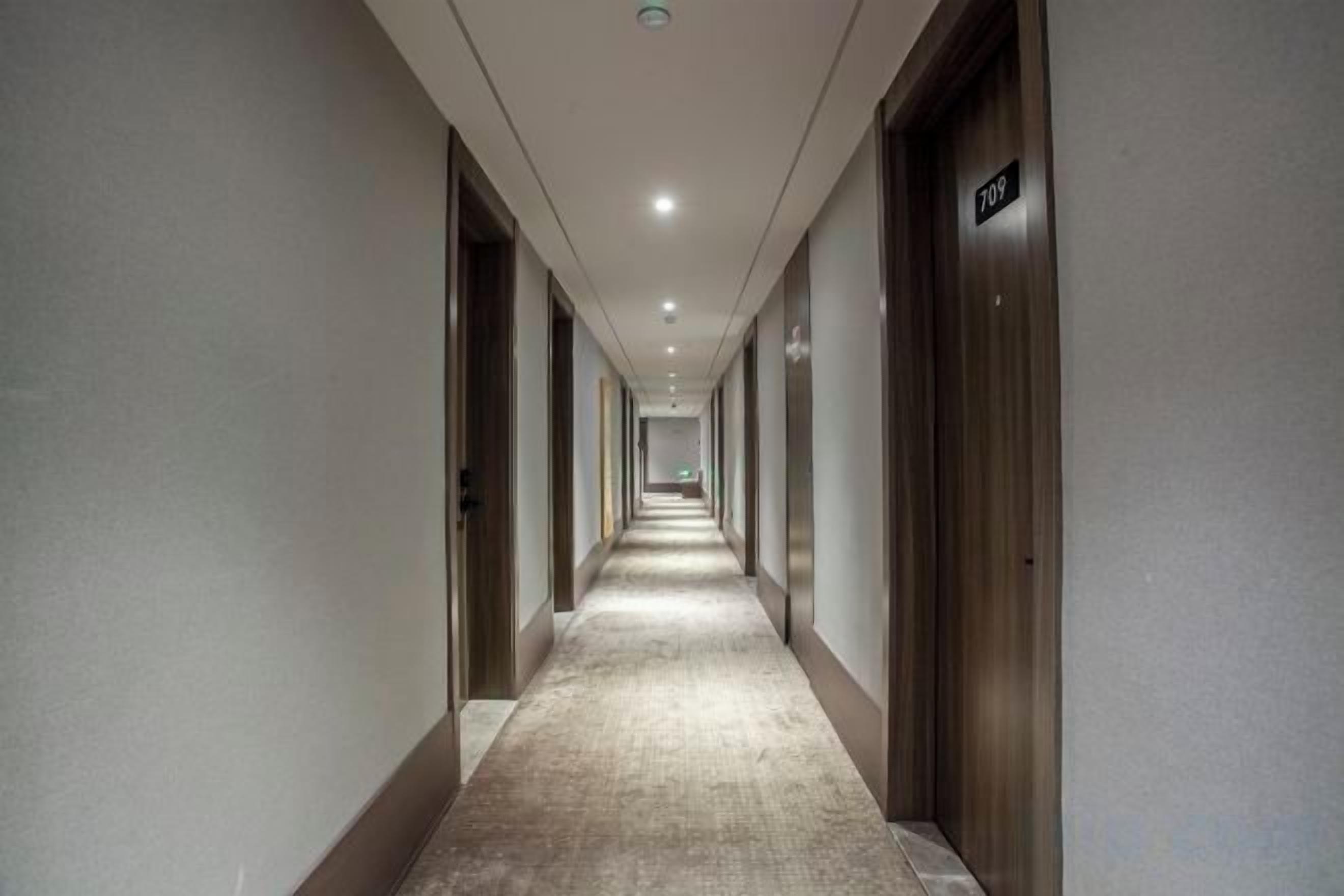 A long, narrow hotel corridor with doors on both sides, a carpeted floor, and a ceiling with recessed lighting.