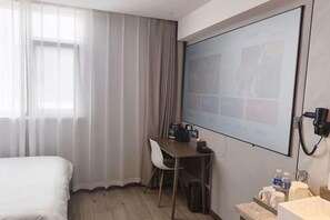 A hotel room with a bed, a desk with a chair, a projector screen, and a window with curtains.