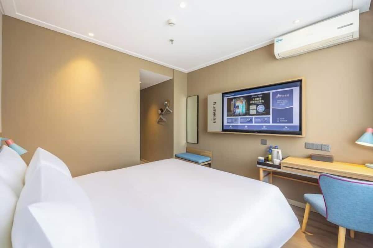 A hotel room with a bed, desk, chair, TV, and an air conditioning unit.