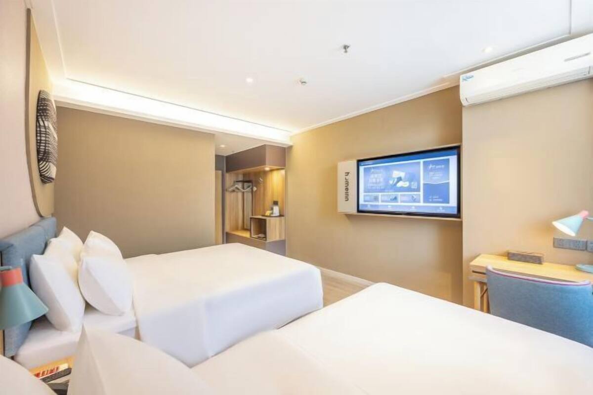A hotel room with two beds, a TV mounted on the wall, a desk with a lamp, and a wardrobe.