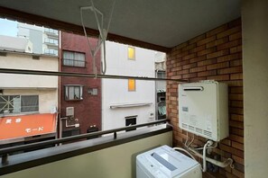 A balcony with a view of adjacent buildings, a brick wall, and a white electrical box. - Japanese Cottage Kenkou (Osaka)