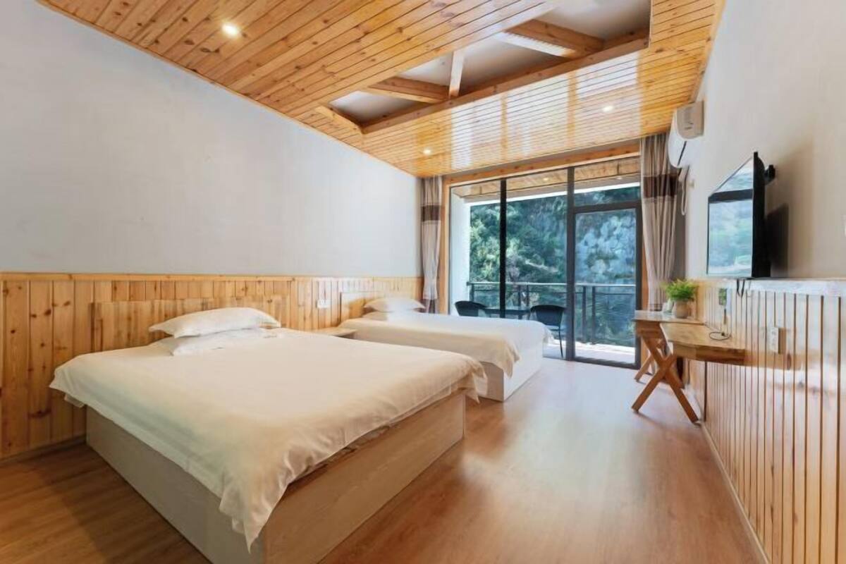 A room with wooden walls and ceiling, two beds, a desk with a chair, a TV, and a large window with a view of trees.