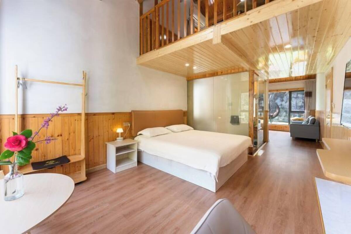 A modern bedroom with a wooden ceiling, a large bed, a bedside table with a lamp, a small table with a vase, and a view of a balcony.