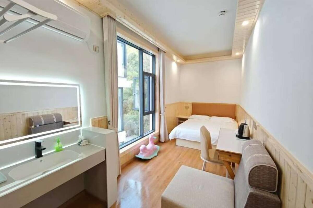 A hotel room with a large window, a bed, a desk, a chair, a small table, a mirror, and a sink.