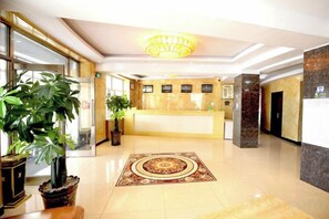 A hotel reception area with a reception desk, potted plants, and a decorative carpet.
