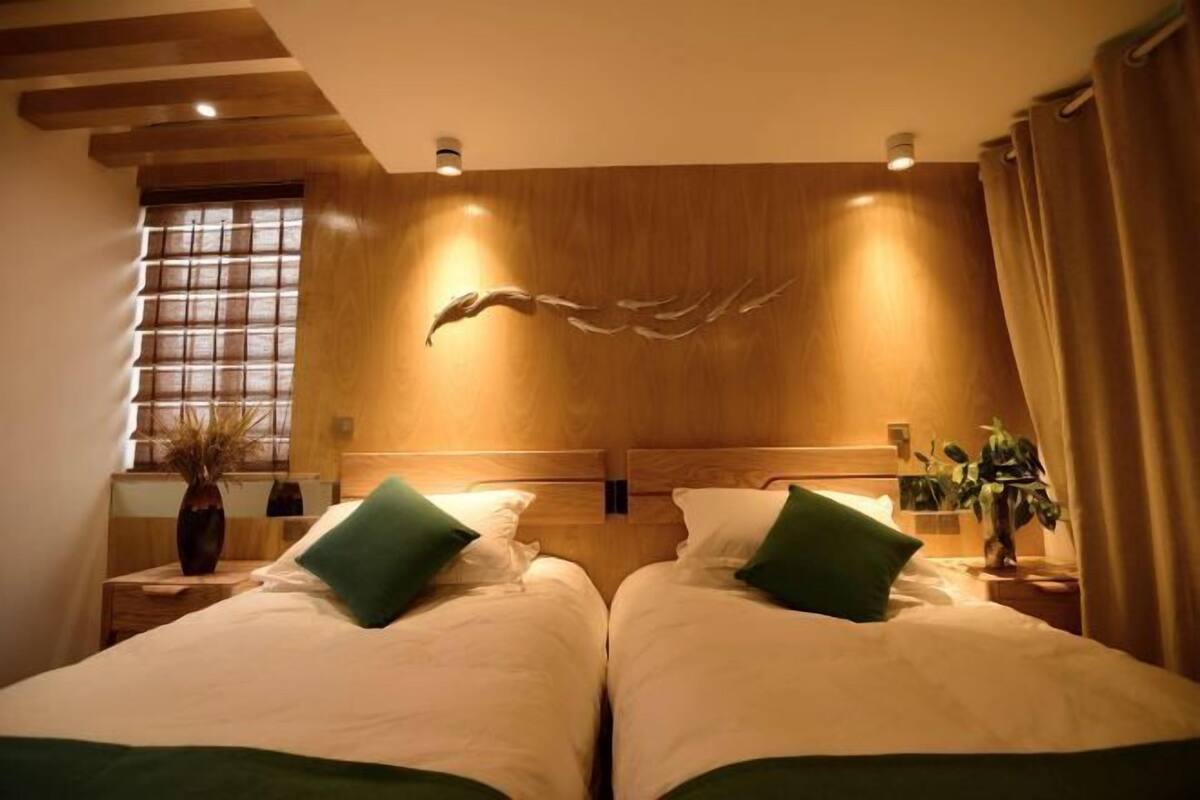 Une chambre avec deux lits, une tête de lit en bois, une fenêtre avec des stores et un luminaire fixé au mur.