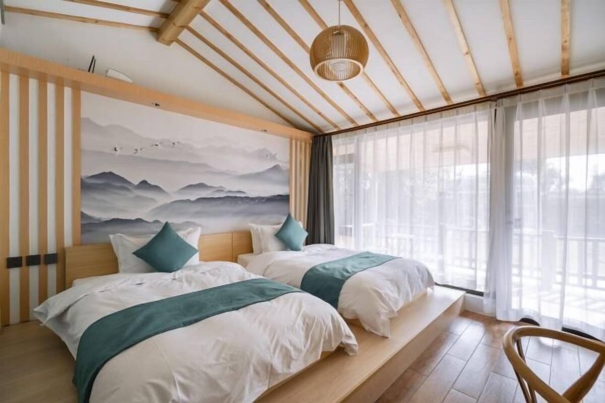 Une chambre d’hôtel avec deux lits, un grand tableau représentant des montagnes, un plafond en bois et une vue sur l’extérieur à travers des rideaux transparents.