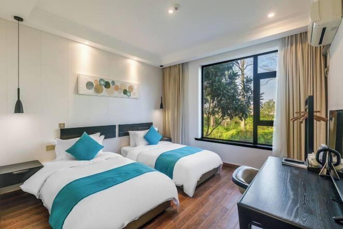 Une chambre d’hôtel avec deux lits, un bureau, une télévision et une grande fenêtre donnant sur un paysage verdoyant.