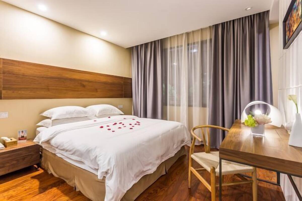 A hotel room with a bed, wooden headboard, a nightstand, a wooden table with a chair, and a window with curtains.