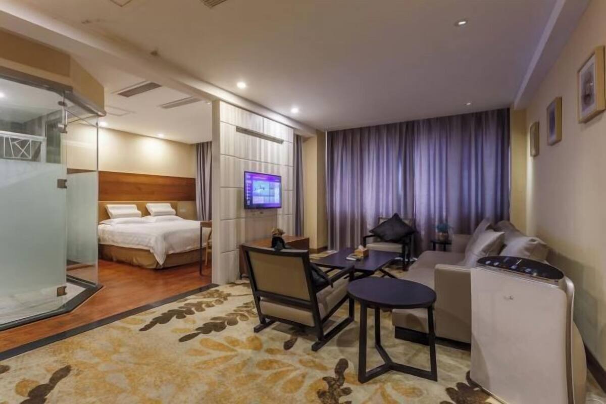 A hotel room with a bed, a desk, a chair, a sofa, a TV, and a shower area.