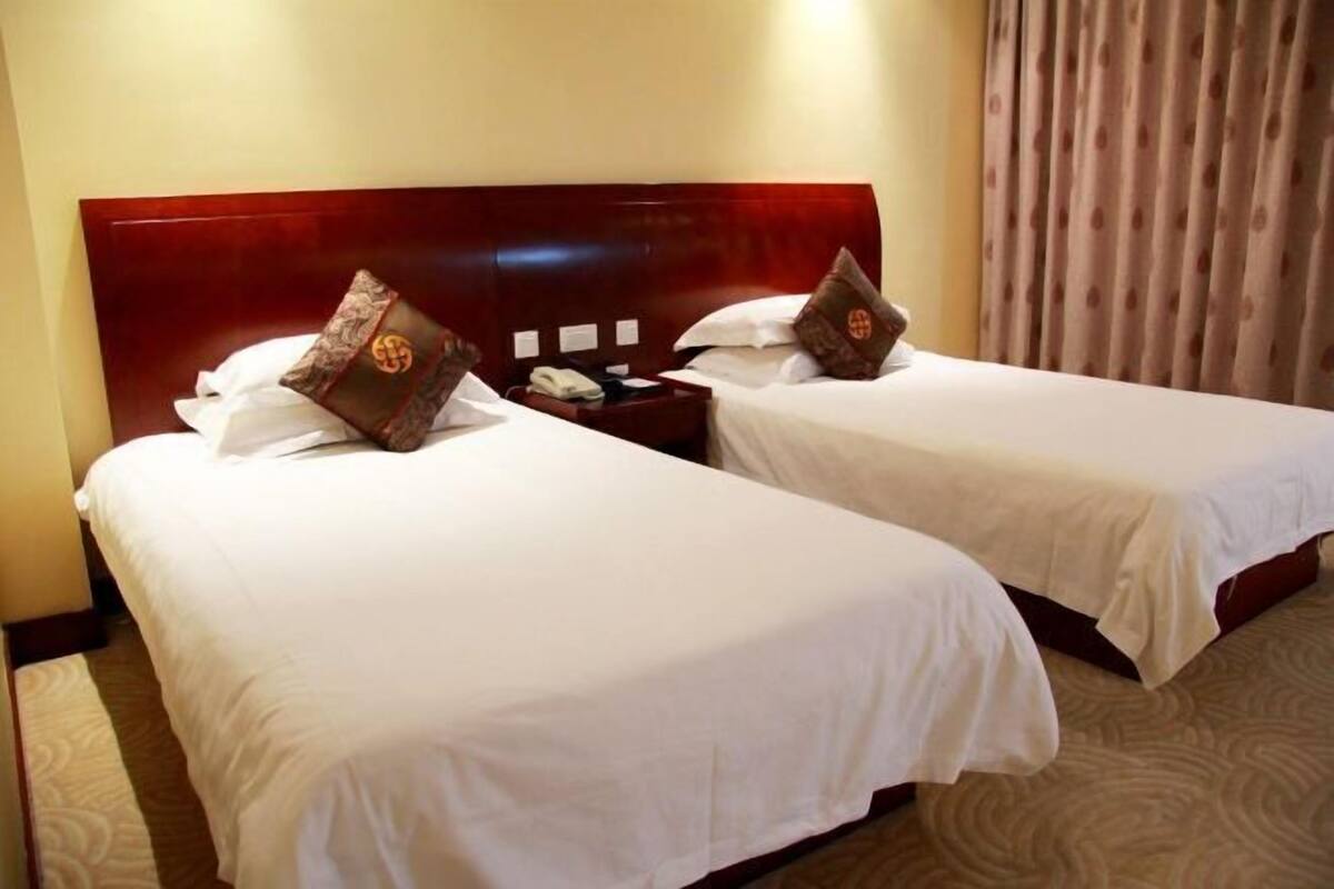 Ein Hotelzimmer mit zwei Betten, jeweils mit weißer Bettwäsche und passenden Kopfteilen. Dazwischen befindet sich ein Nachttisch mit Telefon und einem dekorativen Gegenstand.