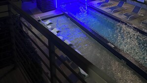 Una piscina amb una estructura iluminada de blau, gandules i un edifici de diversos pisos al fons.