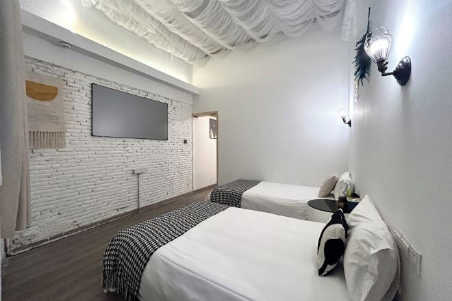 Une chambre d’hôtel avec un mur en briques blanches, un téléviseur à écran plat, deux lits avec du linge de lit blanc, une petite table et un luminaire fixé au mur.