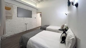 Ein Hotelzimmer mit weißer Ziegelwand, einem Flachbildfernseher, zwei Betten mit weißer Bettwäsche, einem kleinen Tisch und einer wandmontierten Leuchte.